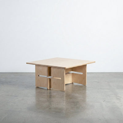 Williamsburg natural wood coffee table with a broad square top and sculptural interlocking panel base with projecting horizontal supports on a concrete floor