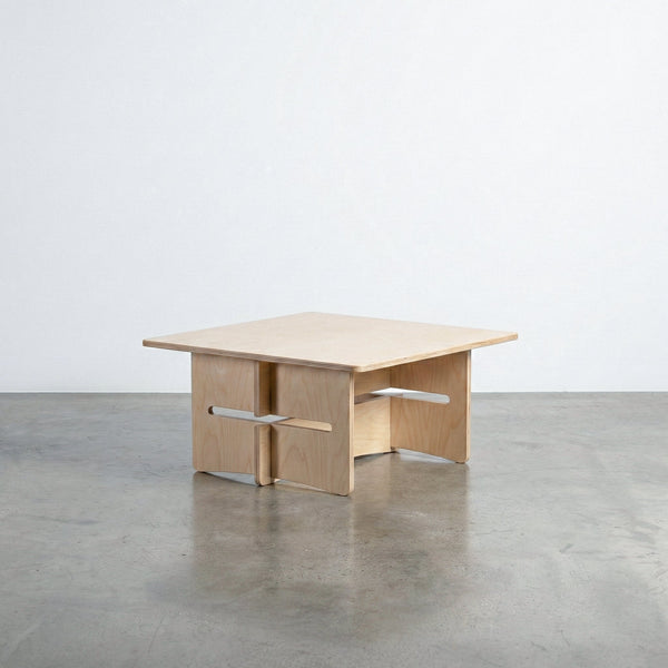 Williamsburg natural wood coffee table with a broad square top and sculptural interlocking panel base with projecting horizontal supports on a concrete floor