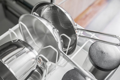 Close-up of the white Yamazaki Home Adjustable Pot Lid Organizer - Steel dividers supporting silver-toned pans, with long handles resting above neighbouring cookware.