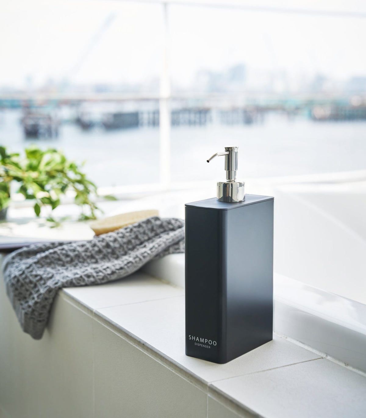 Black Yamazaki Home Rectangle Shower Dispenser - Three Styles labelled SHAMPOO on a white bathtub ledge, with a silver pump, grey textured towel and plant beside a bright window.