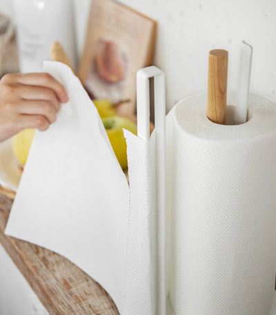 Close-up of a person pulling a paper towel sheet leftward from the Yamazaki Home Paper Towel Holder - Steel + Wood, with the roll resting beside the tall white side rail.