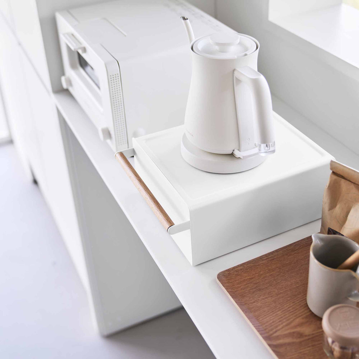 High-angle view of the Yamazaki Home Countertop Rack with Drawer - Steel + Wood beneath a white kettle, showing its flat top, closed drawer and cylindrical wooden handle beside an oven.