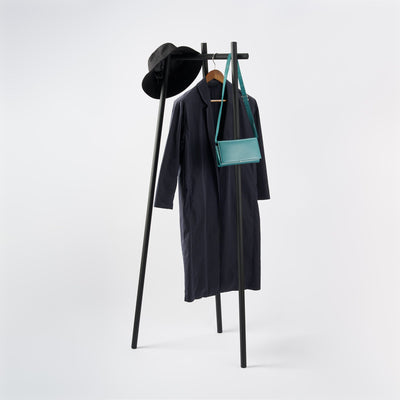 Black Yamazaki Home Tri-Post Coat Rack (60" H) - Steel on a pale background, holding a long dark coat, black hat and small teal shoulder bag.