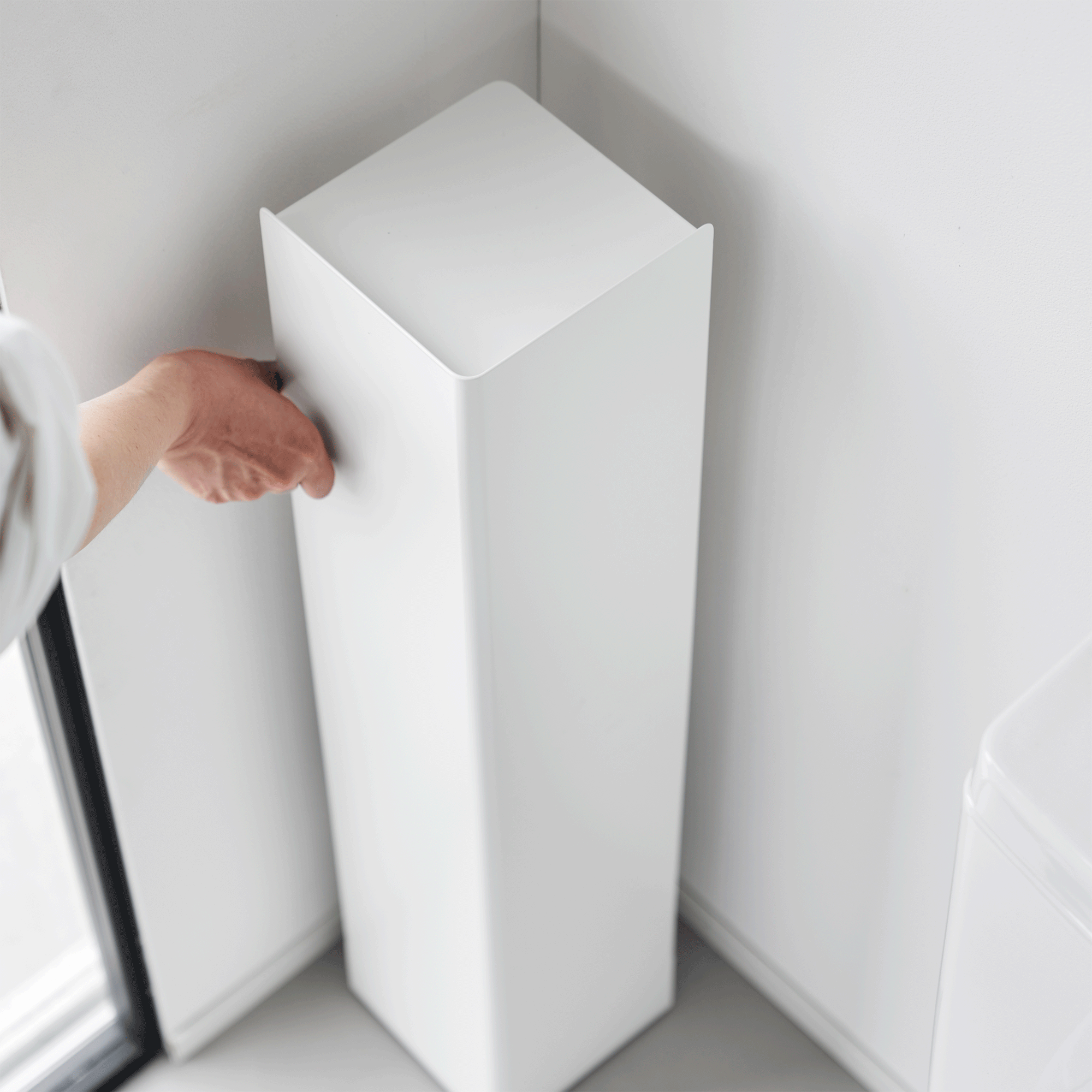 A person turns the white Yamazaki Home Rolling Bathroom Organizer - Steel in a bathroom corner, revealing open shelves holding a toilet brush and cleaning bottle behind its tall solid panels.