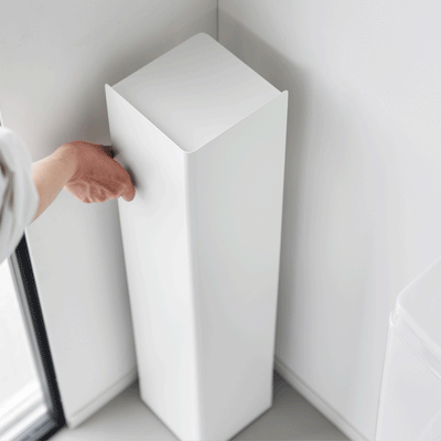 A person turns the white Yamazaki Home Rolling Bathroom Organizer - Steel in a bathroom corner, revealing open shelves holding a toilet brush and cleaning bottle behind its tall solid panels.