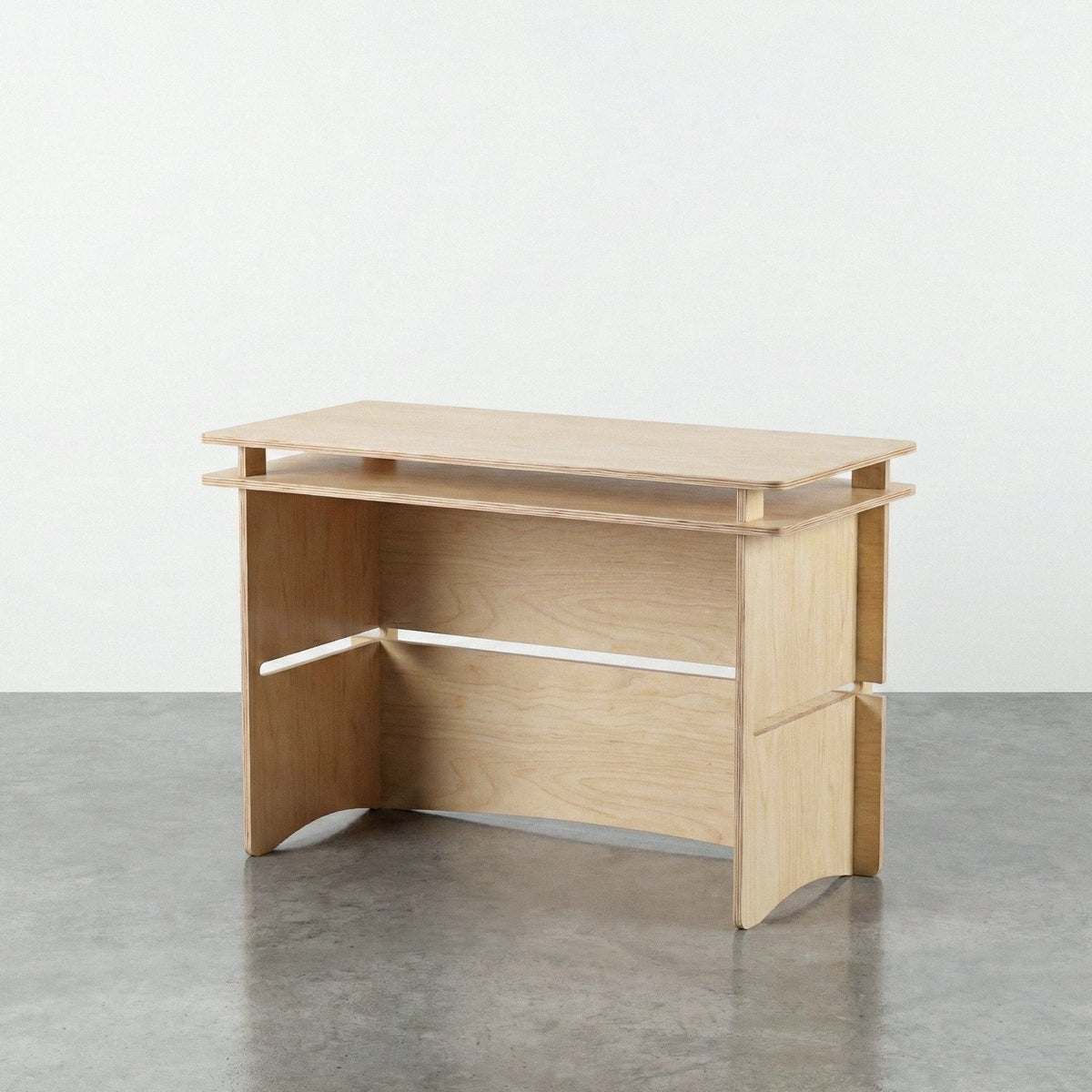 Studio Furnis Williamsburg natural wood desk with a raised desktop shelf, broad panel legs, open knee space and a rear modesty panel on a concrete floor