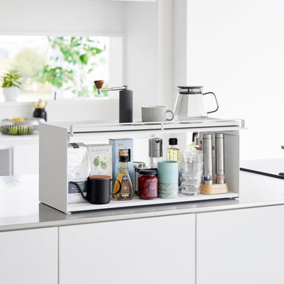White Yamazaki Home Open-Back Countertop Storage Rack - Steel on a kitchen counter with its front panel raised, revealing bottles, jars, drinkware and tall mills; a coffee grinder, mug and glass carafe sit on top.