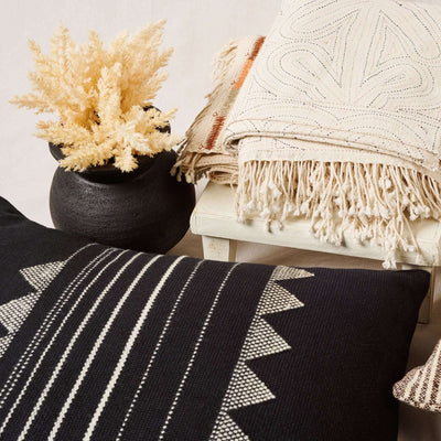 Kora Black pillow styled with a black vase of dried grasses and layered cream fringed throws