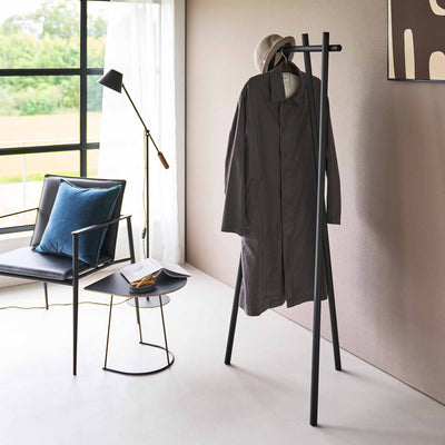 Black Yamazaki Home Tri-Post Coat Rack (60" H) - Steel supporting a long dark coat and neutral-coloured hat beside a beige wall, with daylight entering through a nearby window.