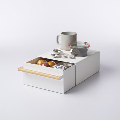 White Yamazaki Home Countertop Rack with Drawer - Steel + Wood with its wood-handled drawer partly open, holding coffee capsules and small containers; a mug, glass jar and measuring spoons rest on top.