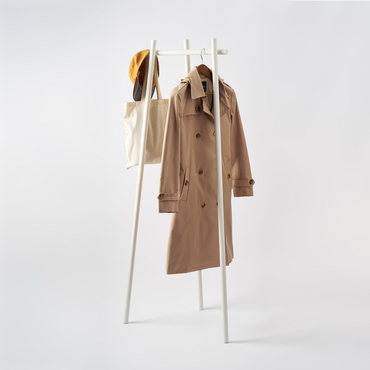 White Yamazaki Home Tri-Post Coat Rack (60" H) - Steel on a pale background, with three slender splayed legs and a horizontal hanging rail holding a tan trench coat, cream tote and mustard-coloured hat.