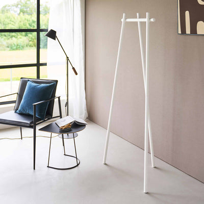 Empty white Yamazaki Home Tri-Post Coat Rack (60" H) - Steel beside a beige wall, showing its three tall posts, splayed feet and short horizontal rail near a window and lounge chair.