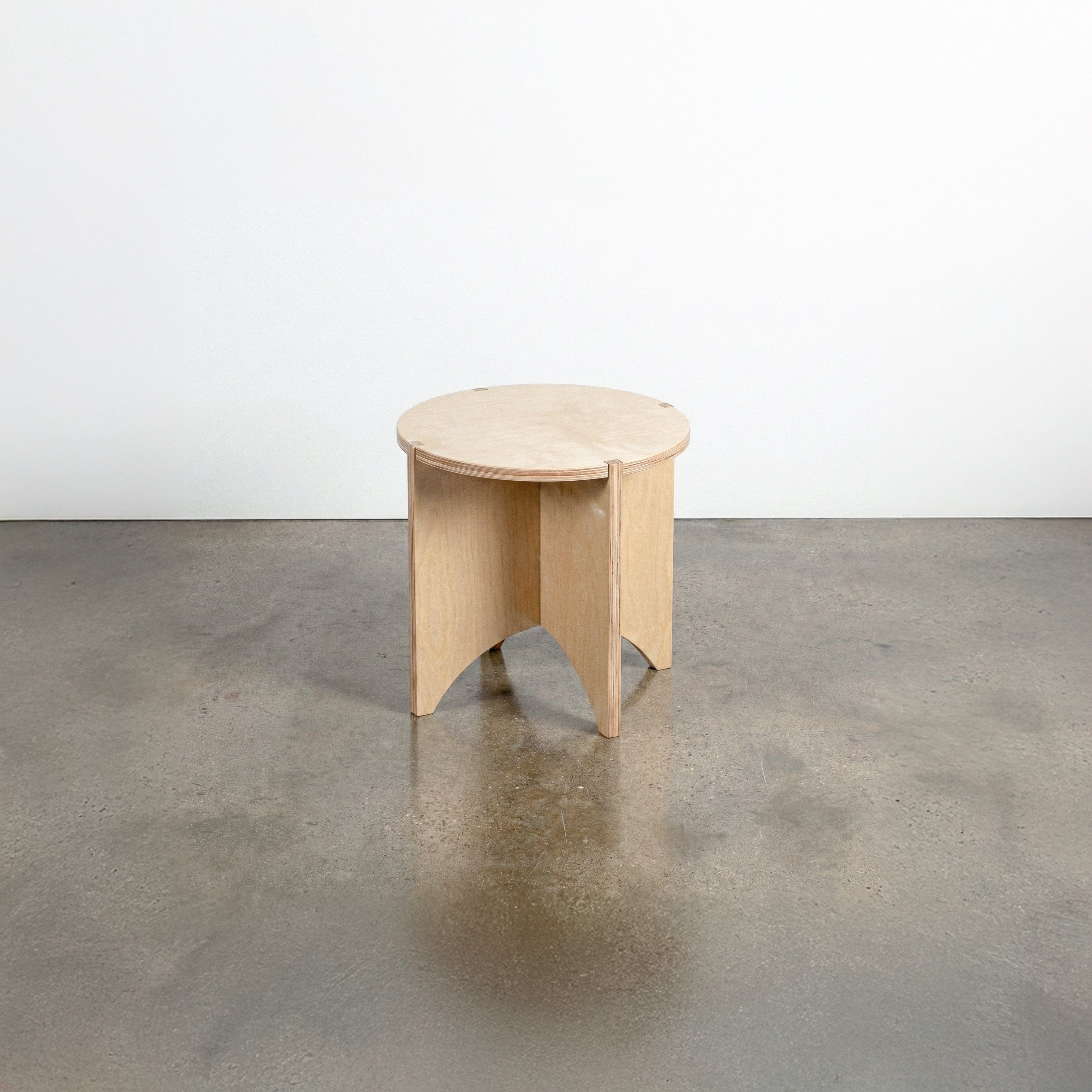 Studio Furnis YOYO natural wood end table with a round top and sculptural intersecting panel legs, displayed on a polished concrete floor