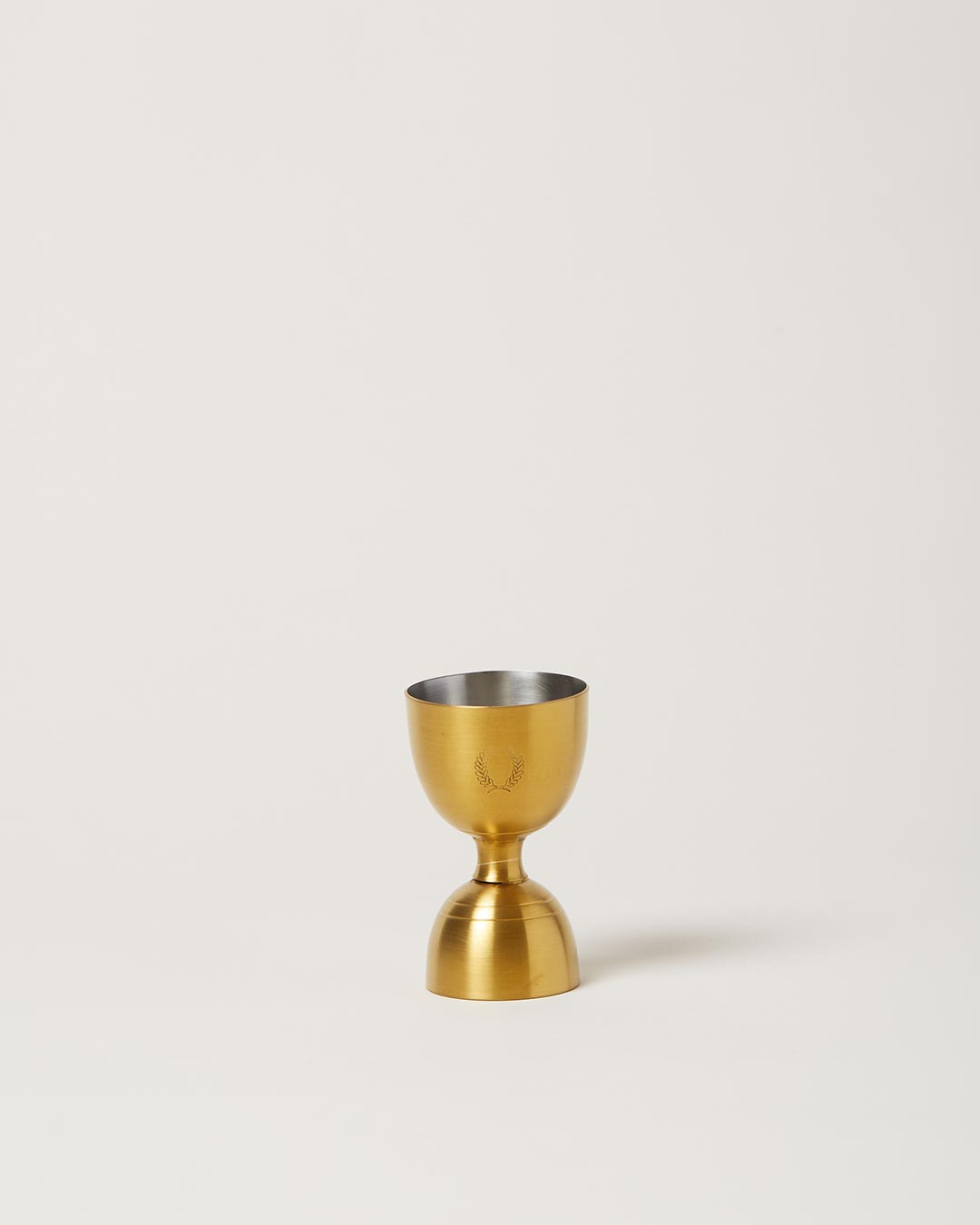 Double-sided brushed-gold jigger standing upright, with a smaller upper cup and larger lower cup lined in silver.