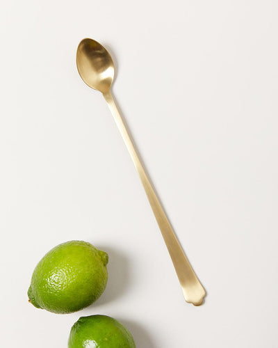 Long brushed-gold bar spoon displayed beside two whole limes, showing its small oval bowl and slender handle.