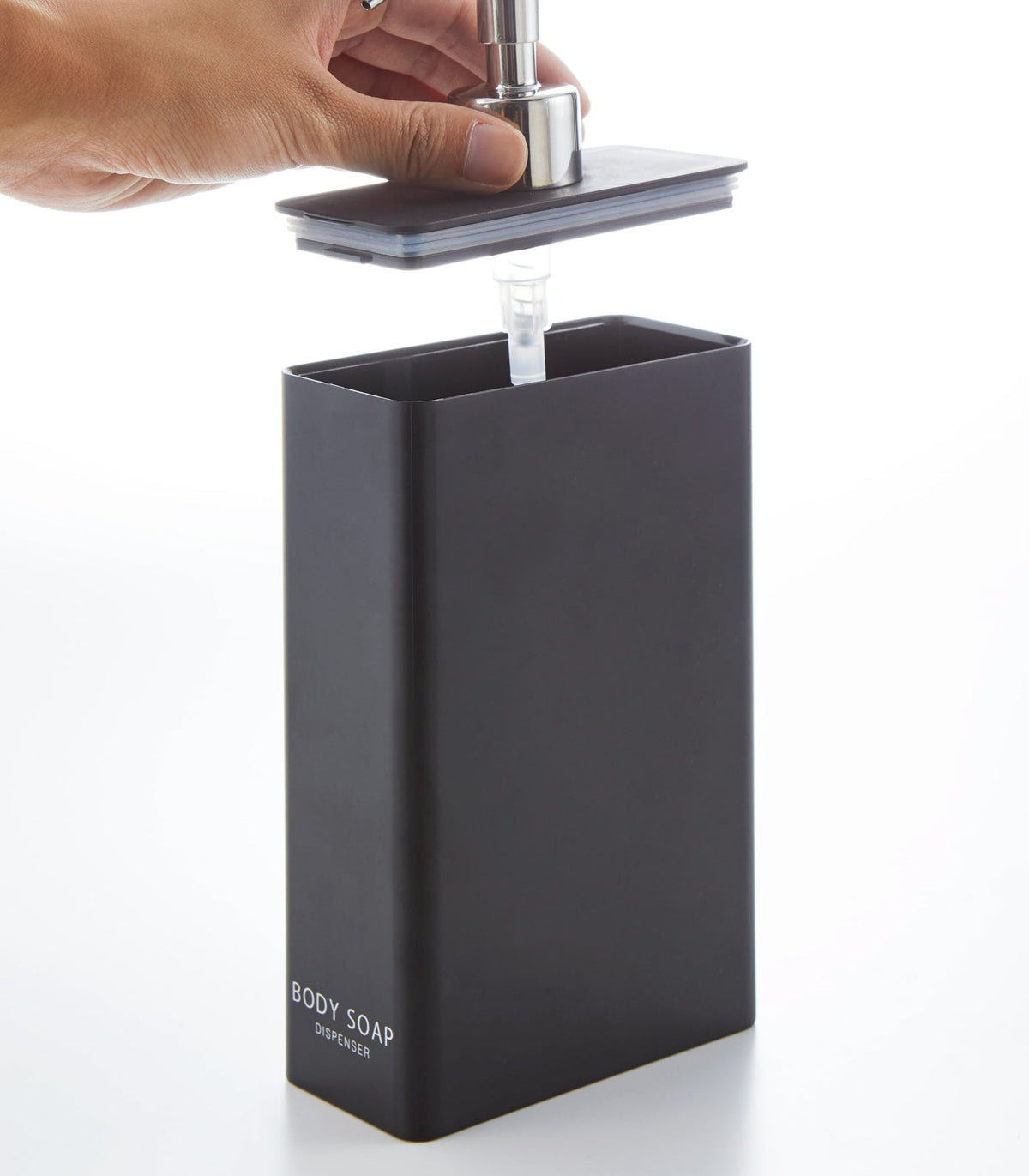 A hand raises the pump-and-lid assembly from the black Yamazaki Home Rectangle Shower Dispenser - Three Styles labelled BODY SOAP, showing the clear tube beneath.
