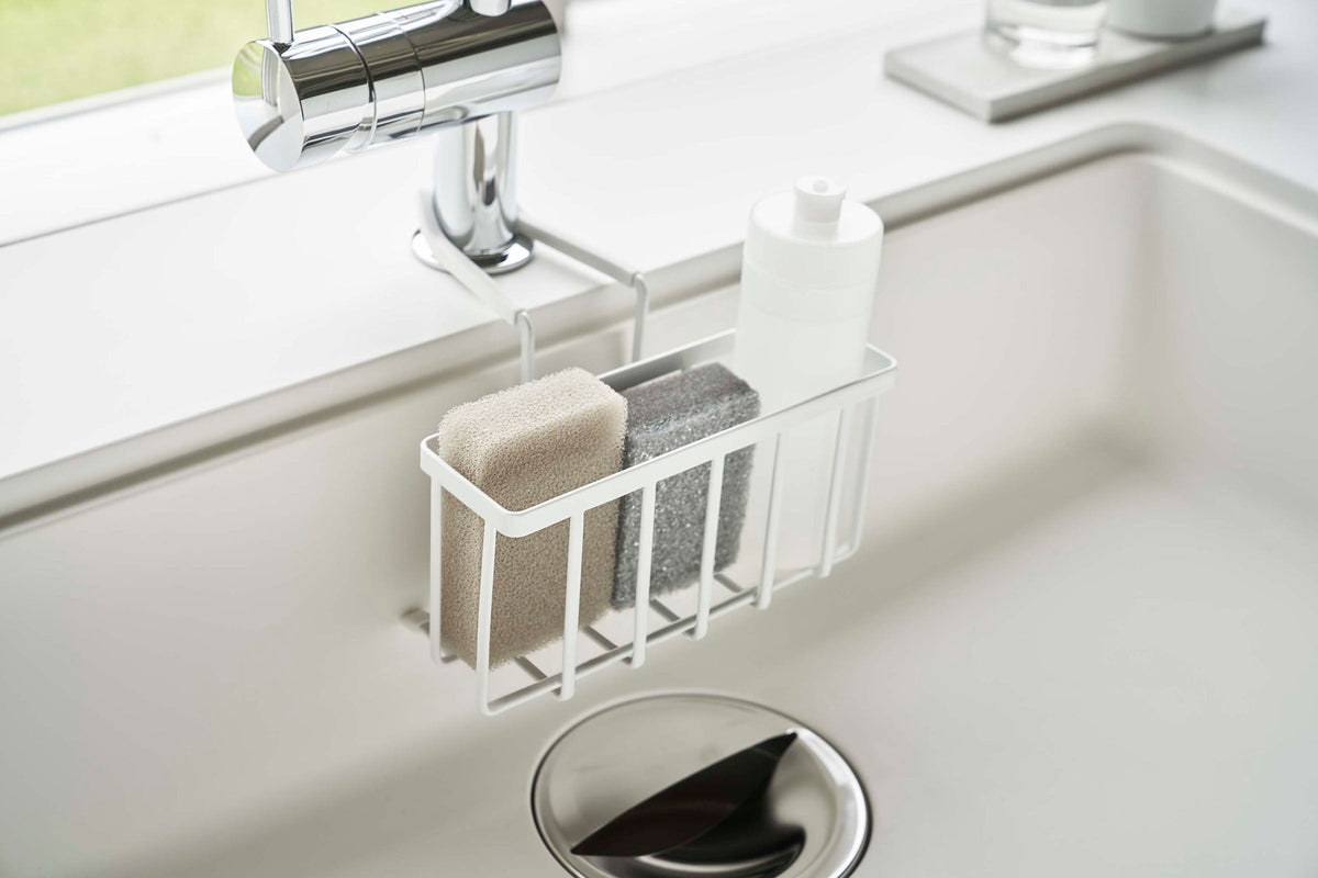 Wider view of the white Yamazaki Home Faucet-Hanging Sponge Caddy - Steel beneath a chrome faucet, holding two sponges and a white bottle above the circular drain.