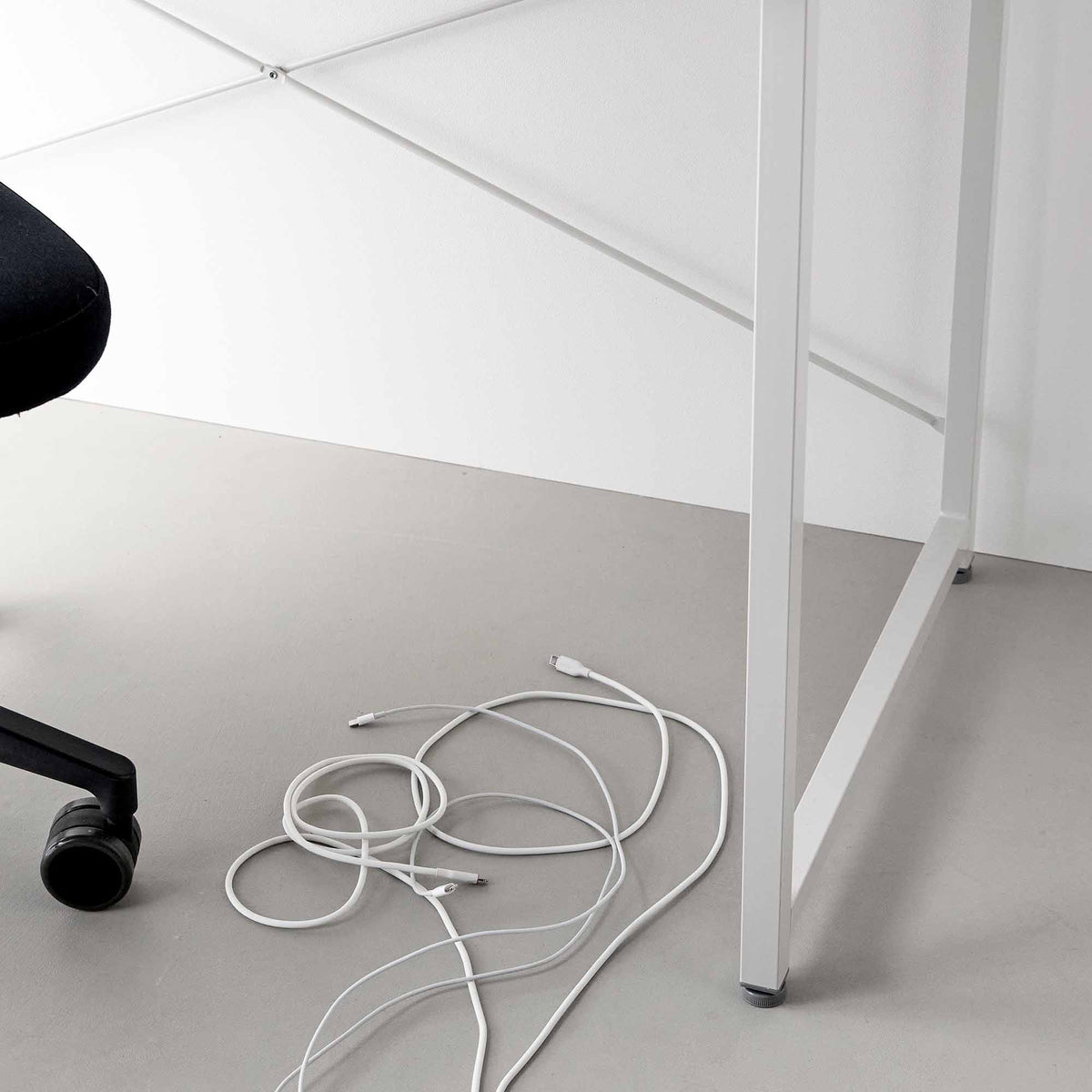 Loose white charging cables coiled on the grey floor beneath a white desk, illustrating the cable arrangement before using Yamazaki Home Magnetic Cable Holders With Adhesive Steel Bases (Set of 5) - Silicone.