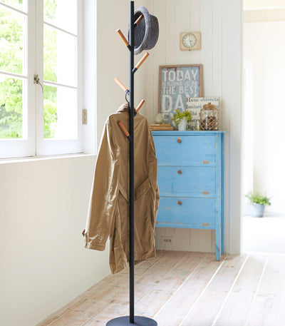 Black Yamazaki Home Coat Rack (70" H) - Steel holding a tan coat and dark hat beside a bright window and blue chest of drawers.
