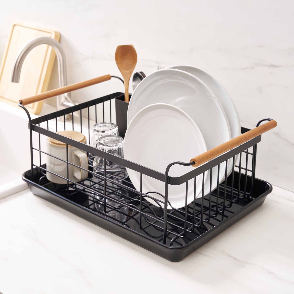 Opposite angled view of the Yamazaki Home Dish Rack - Steel + Wood on a pale counter, showing wooden side handles, upright plates, inverted glasses and utensils beside the faucet.