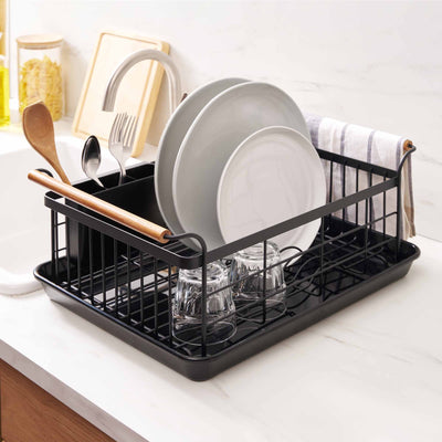Yamazaki Home Dish Rack - Steel + Wood beside a kitchen sink, viewed diagonally with pale plates standing in the black wire slots, glasses below and a striped towel draped over the rear edge.