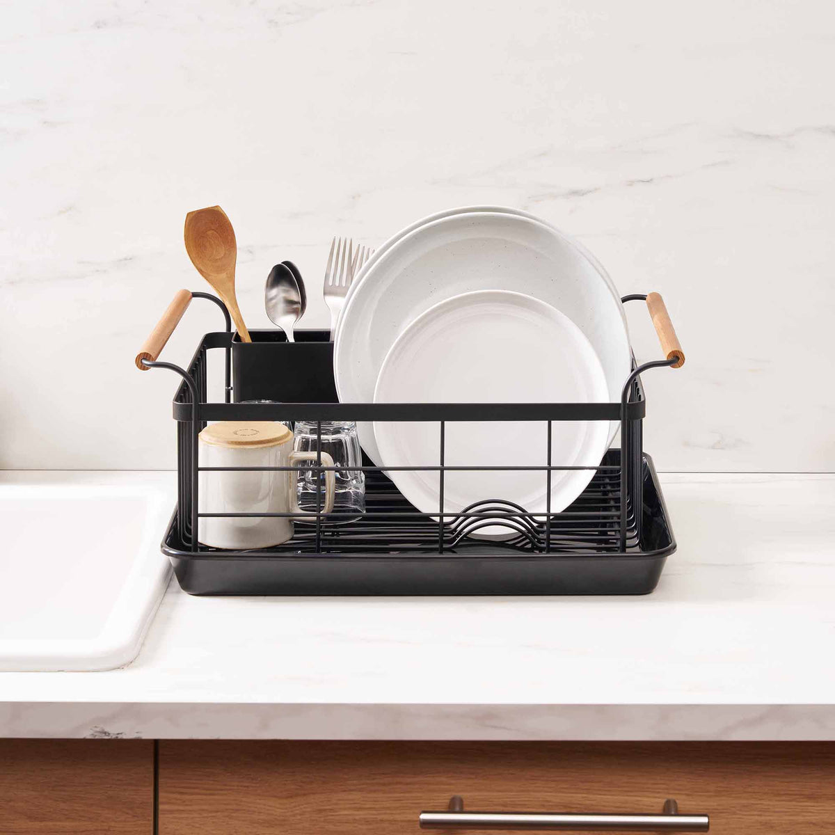 Front view of the black Yamazaki Home Dish Rack - Steel + Wood beside a sink, with pale plates arranged upright, utensils at the back and the basket resting inside a shallow rectangular drip tray.