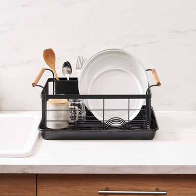 Front view of the black Yamazaki Home Dish Rack - Steel + Wood beside a sink, with pale plates arranged upright, utensils at the back and the basket resting inside a shallow rectangular drip tray.