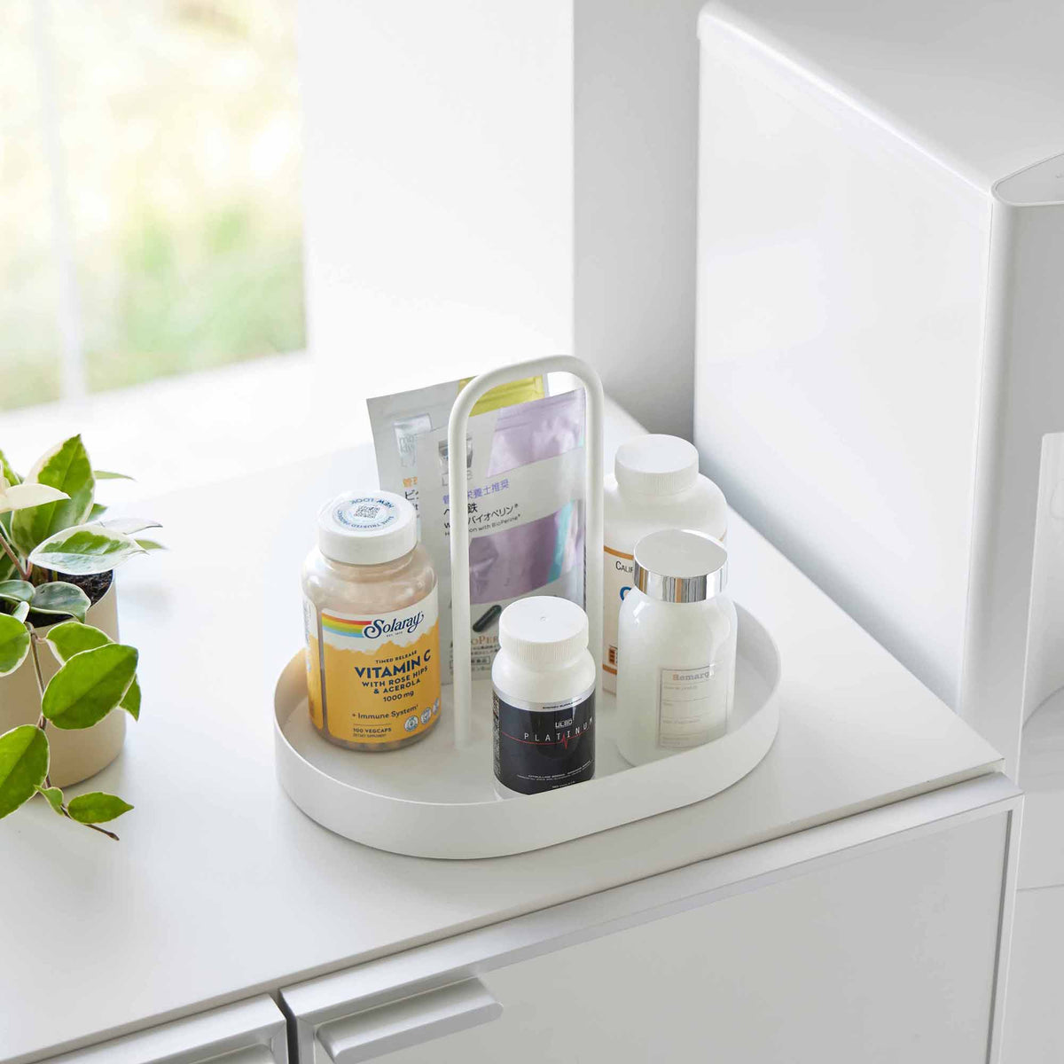 Angled view of the white Yamazaki Home Vanity Tray with Handle - Steel holding bottles and packets on a cabinet beside a window and small potted plant.