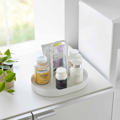 Angled view of the white Yamazaki Home Vanity Tray with Handle - Steel holding bottles and packets on a cabinet beside a window and small potted plant.