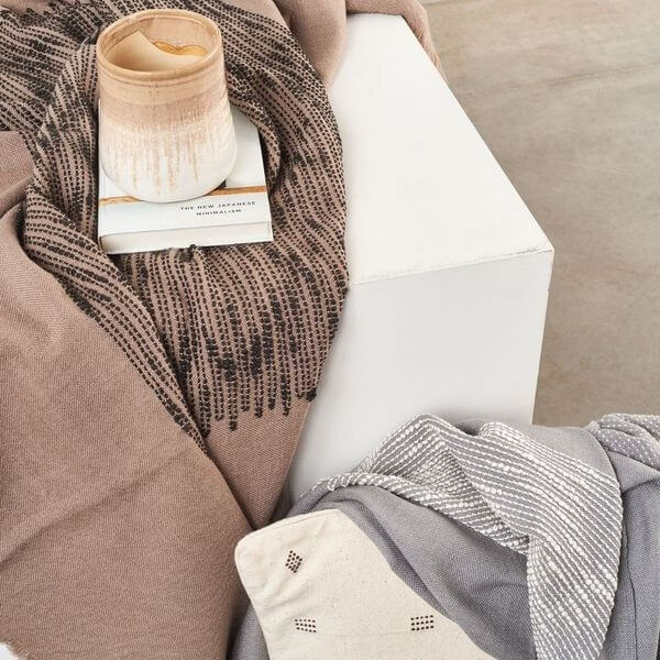 Flo Brown merino throw styled over a white side table with books and a candle beside a folded gray textile