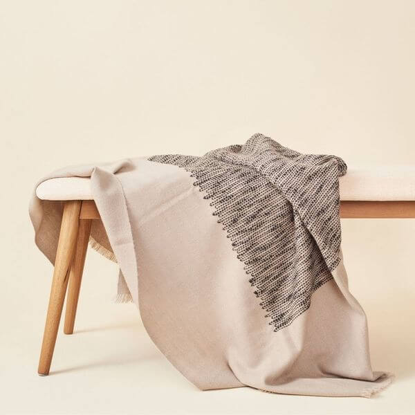 Flo Brown handloom throw draped over a pale wood bench, showing its warm brown field, dark woven pattern and fringed edge