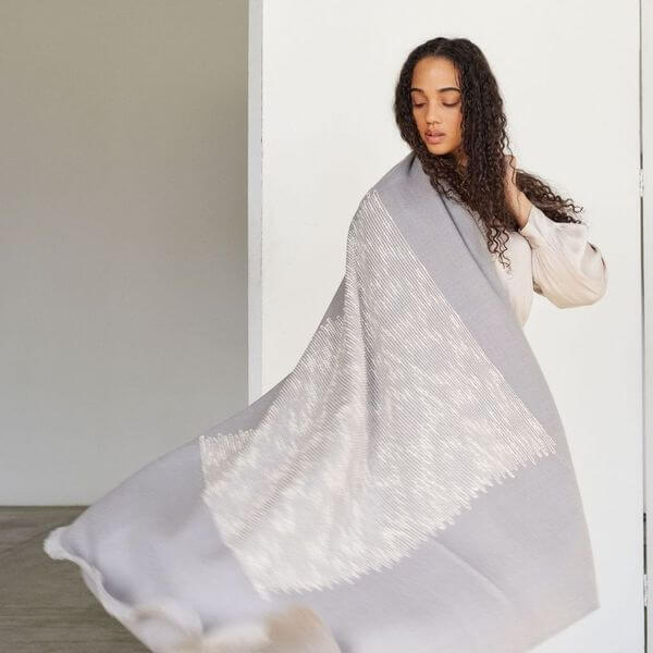 Woman holding the Flo Gray handloom throw open to display its full size, pale gray color and large white woven design