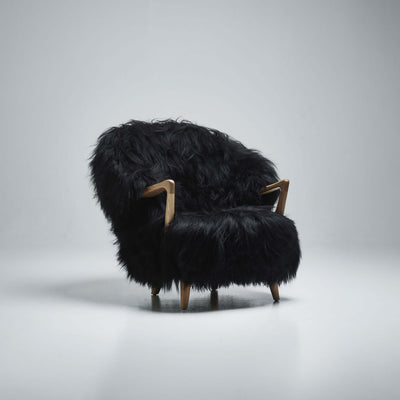 Three-quarter view of the black Eikund Fluffy Lounge Chair in a gray studio, showing its rounded back, thick shaggy seat, angular wood armrests, and short legs.