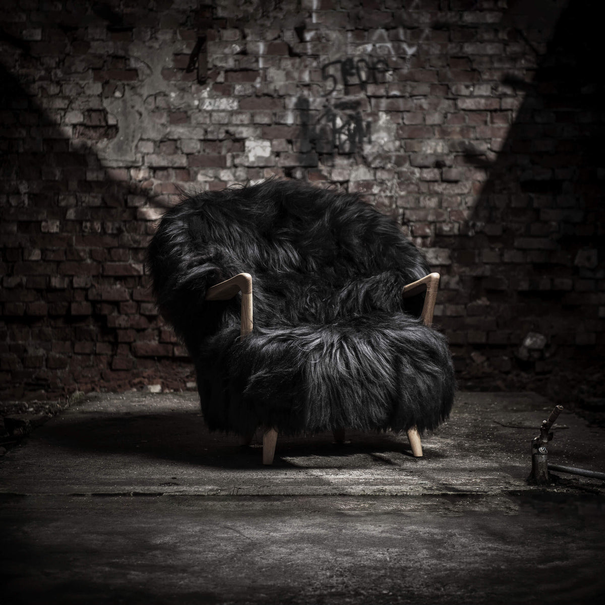 Front three-quarter view of the black Eikund Fluffy Lounge Chair against a distressed brick wall, showing its broad curved back, shaggy seat, exposed wood arms, and short legs on a worn floor.