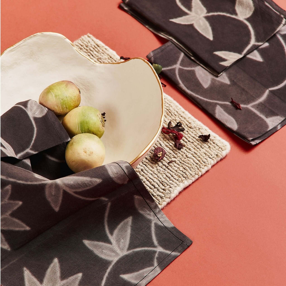 Overhead table setting with Folio Black leaf-print napkins, a white plate of green apples and a woven placemat