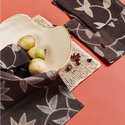 Overhead table setting with Folio Black leaf-print napkins, a white plate of green apples and a woven placemat