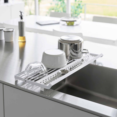 White Yamazaki Home Slim Over-the-Sink Dish Drying Rack - Steel used flat across a sink, holding an inverted saucepan, white bowl, clear glass bowl and utensils.