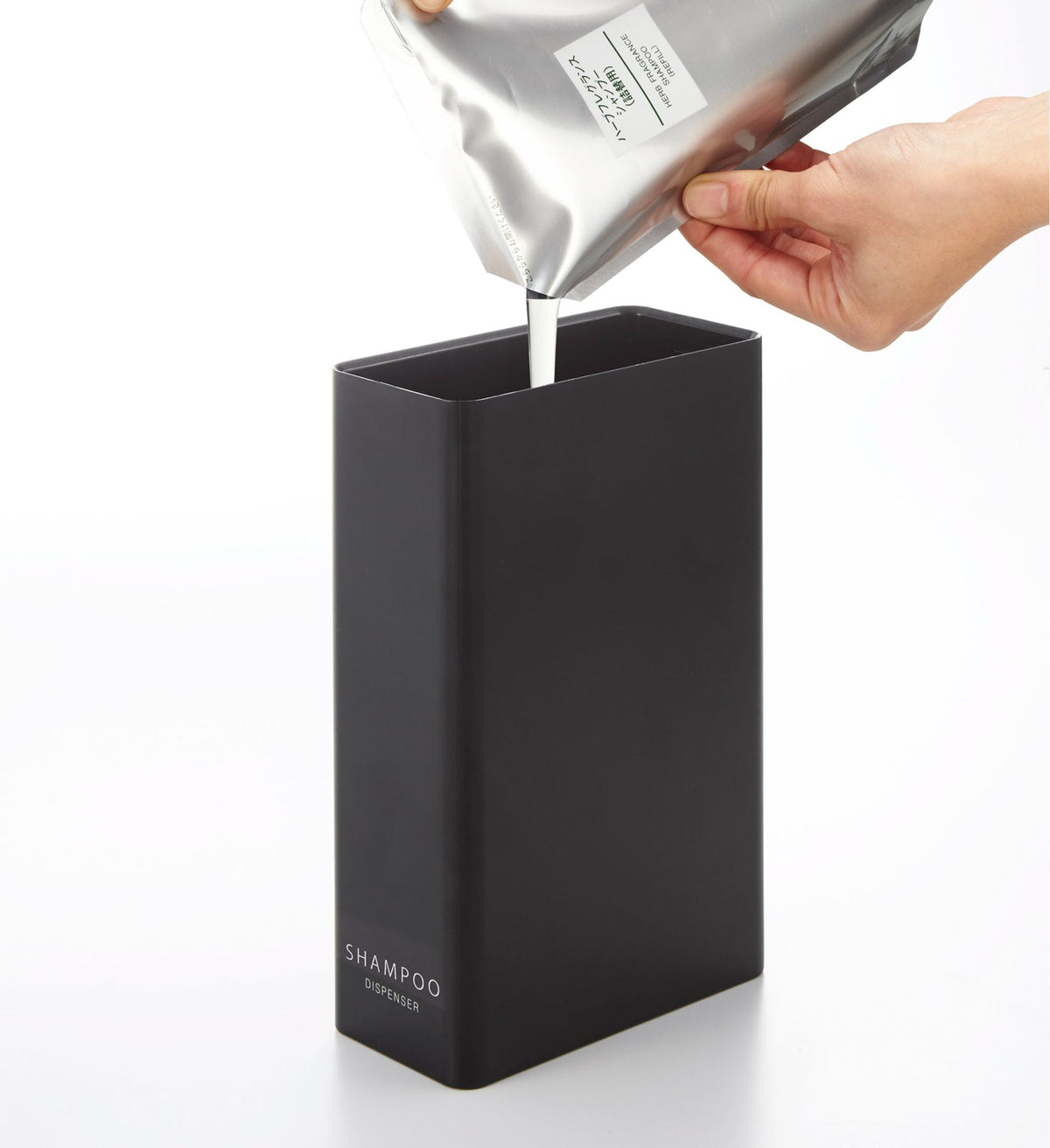 Liquid pours from a silver refill pouch into the open black Yamazaki Home Rectangle Shower Dispenser - Three Styles labelled SHAMPOO.