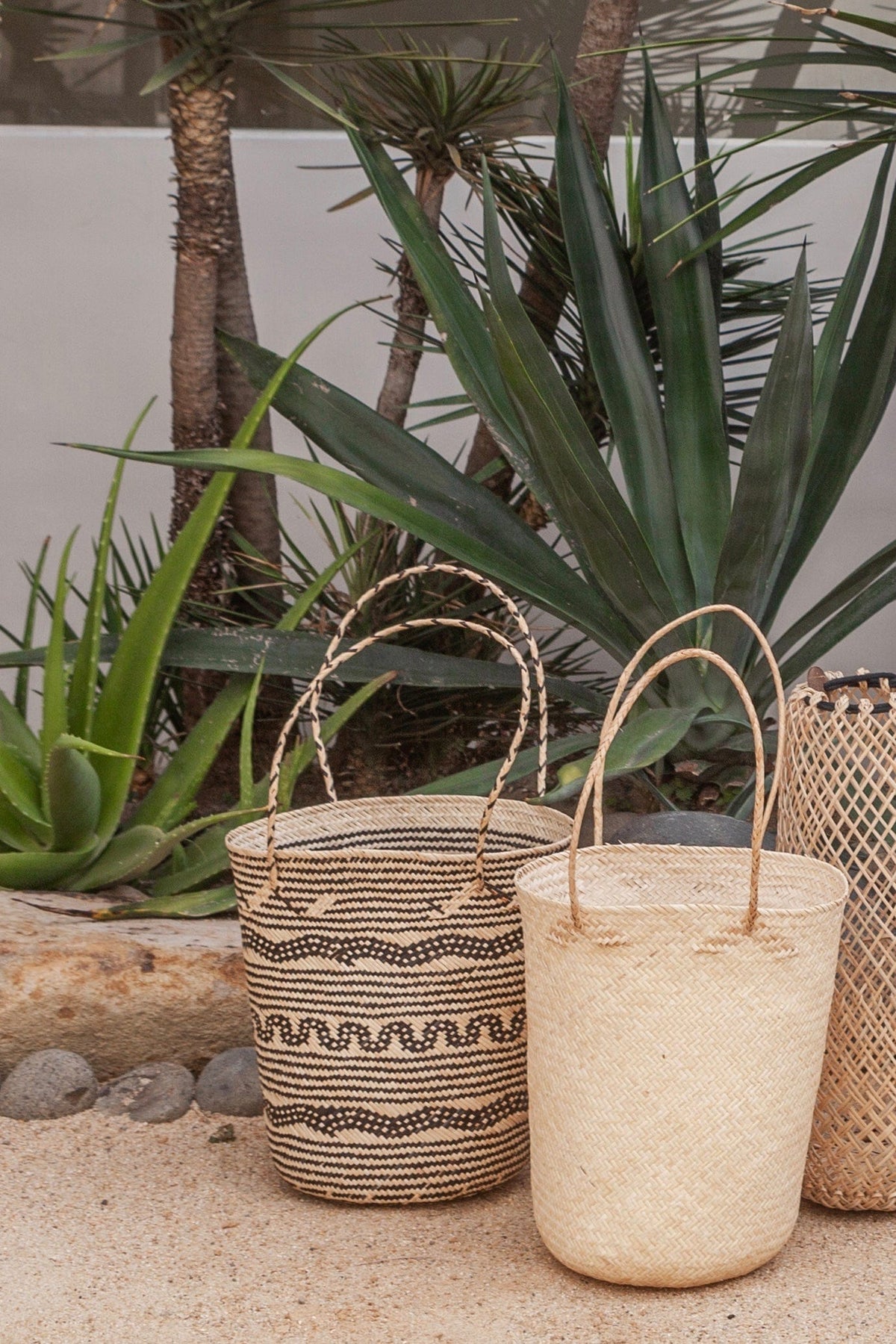 Village Thrive Praya Day Bag, natural and black rattan, three bags displayed among potted plants.