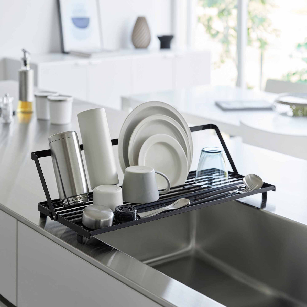 Black Yamazaki Home Slim Over-the-Sink Dish Drying Rack - Steel spanning a stainless-steel sink, holding plates, tall drinkware, mugs and utensils beneath its raised support rail.
