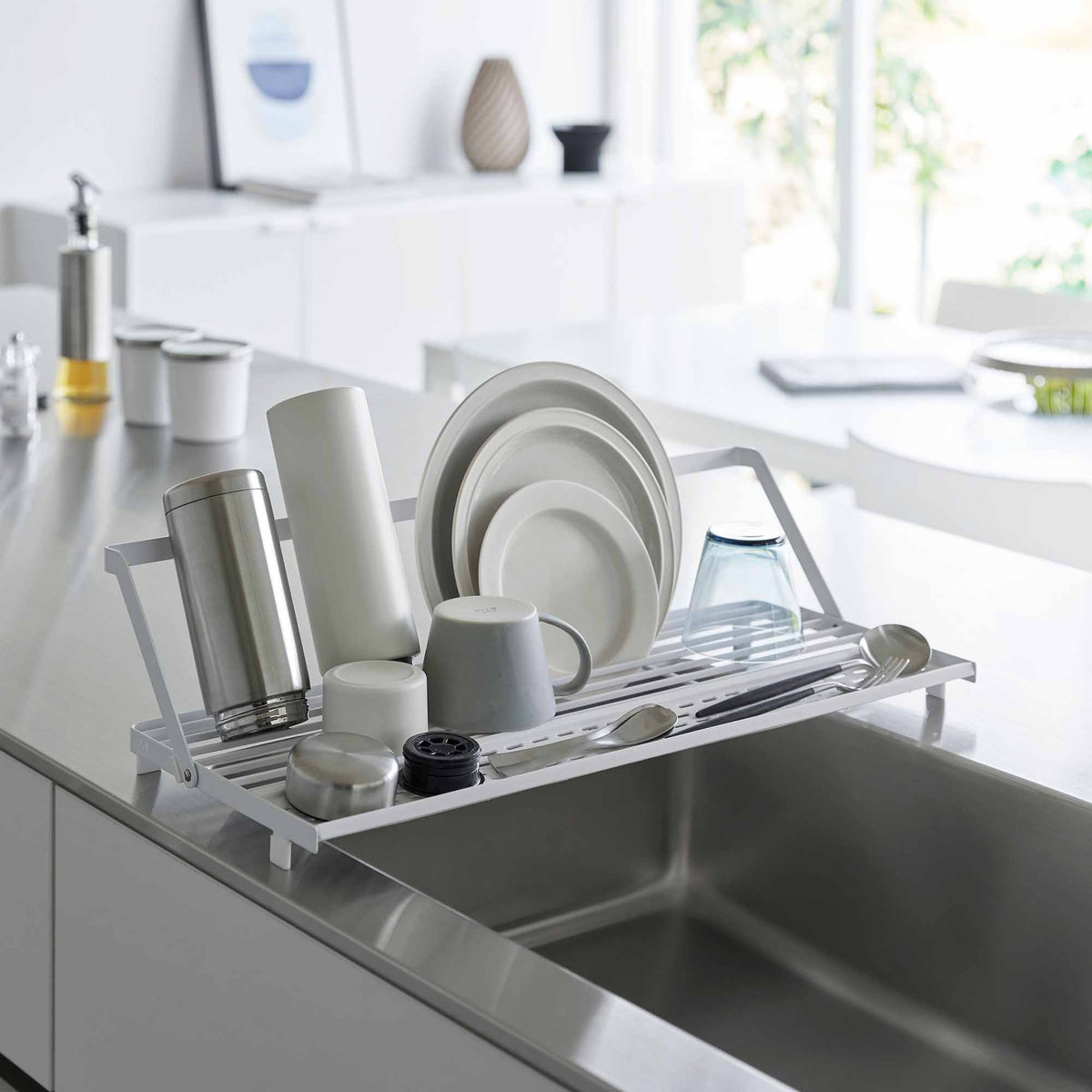 White Yamazaki Home Slim Over-the-Sink Dish Drying Rack - Steel spanning a stainless-steel sink, with upright plates, a tall tumbler, mugs and a blue glass arranged across its narrow frame.