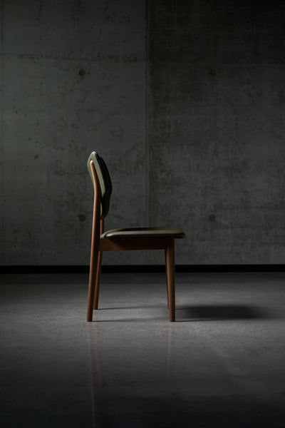 Side profile of the dark green Eikund Veng Dining Chair, showing the reclined backrest, thin padded seat, and slender angled wood frame against a concrete wall.