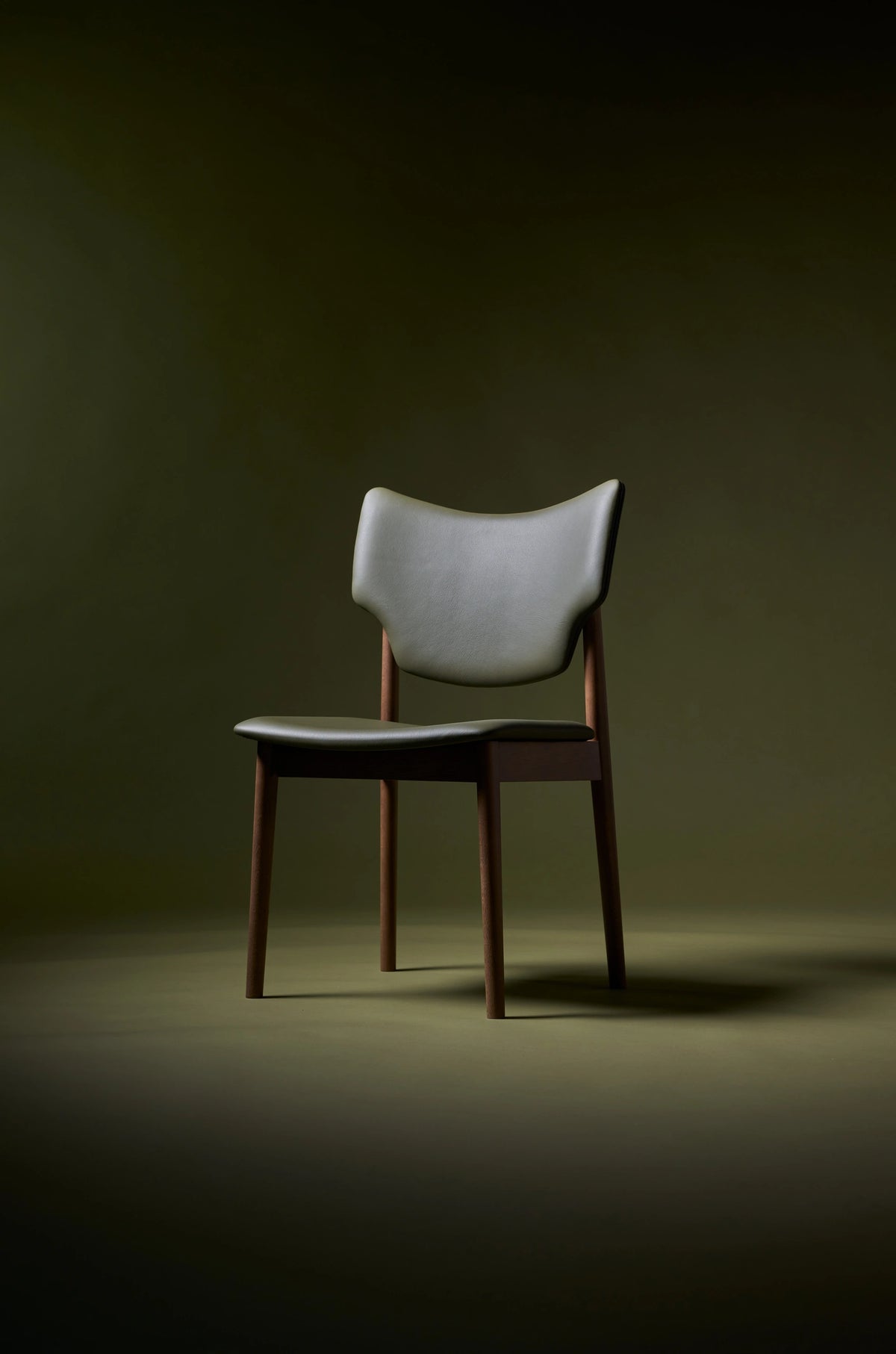 Three-quarter front view of the Eikund Veng Dining Chair under dramatic lighting against an olive background, showing a gray-green upholstered back, dark seat, and brown wood frame.