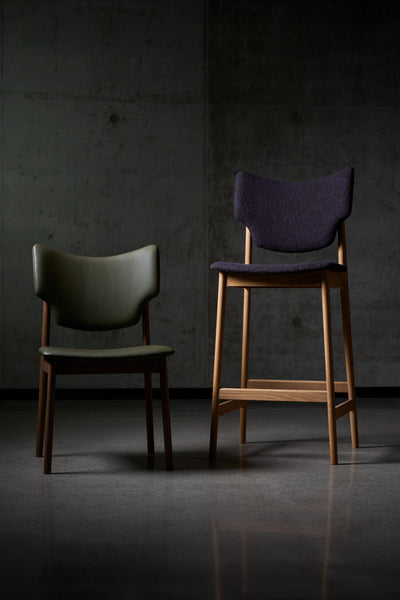 Dark purple Eikund Veng Bar Stool beside a lower chair with a dark green backrest, showing their similar curved back shapes and contrasting heights against a concrete wall.