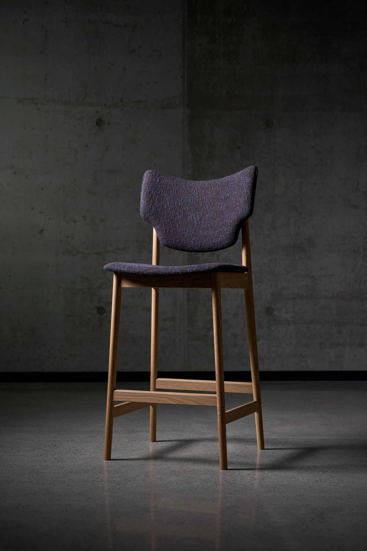 Three-quarter view of the Eikund Veng Bar Stool with dark purple textured upholstery and a warm brown wood frame, standing against a concrete wall on a dark tiled floor.