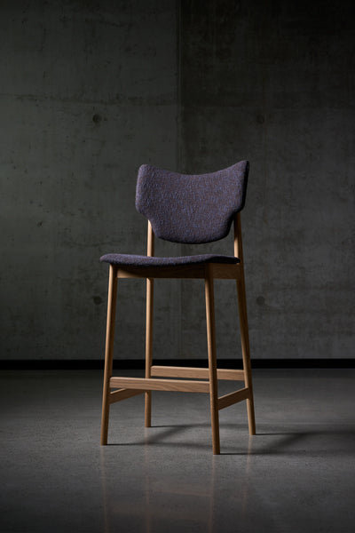 Three-quarter view of the Eikund Veng Bar Stool with dark purple textured upholstery and a warm brown wood frame, standing against a concrete wall on a dark tiled floor.