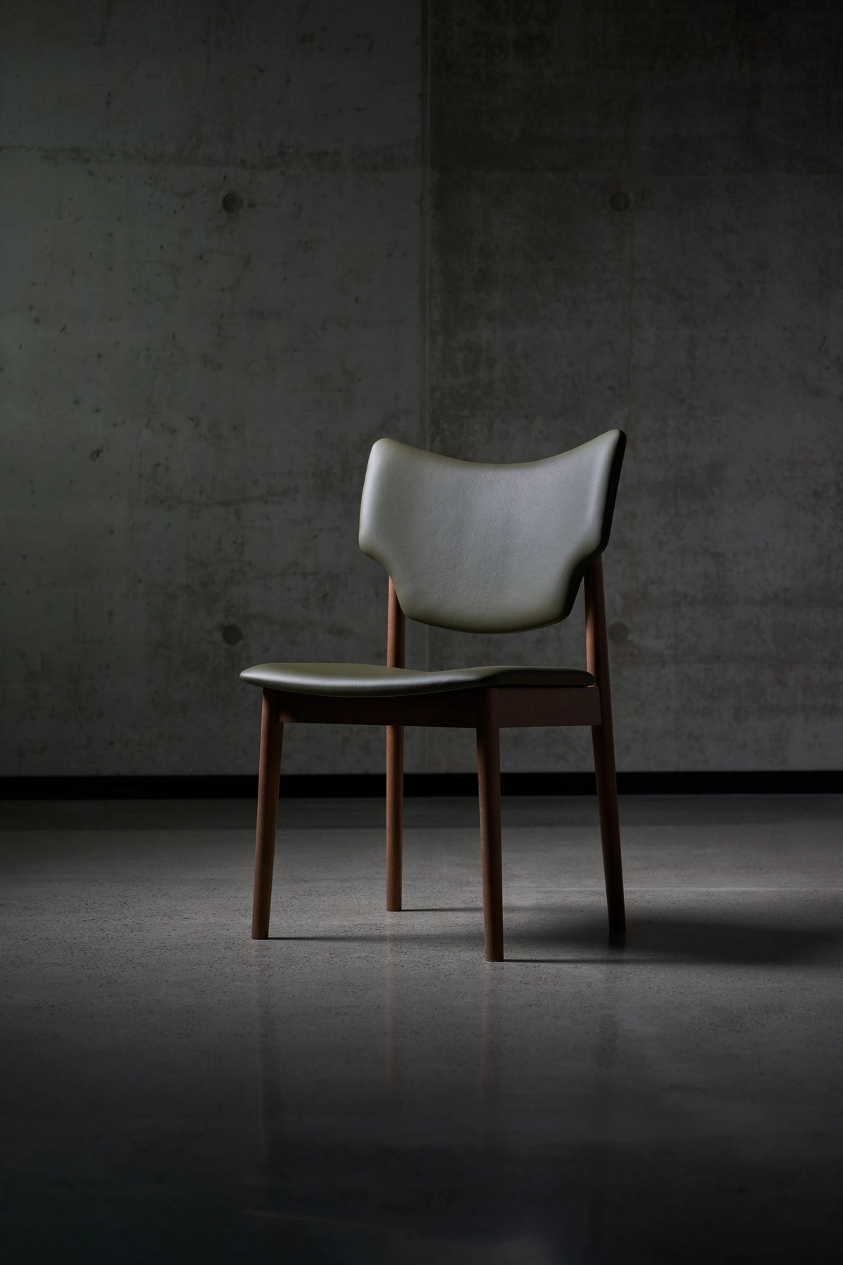 Angled front view of the gray-green Eikund Veng Dining Chair against a concrete wall, showing its sculpted backrest, separate seat, and tapered brown wood legs.