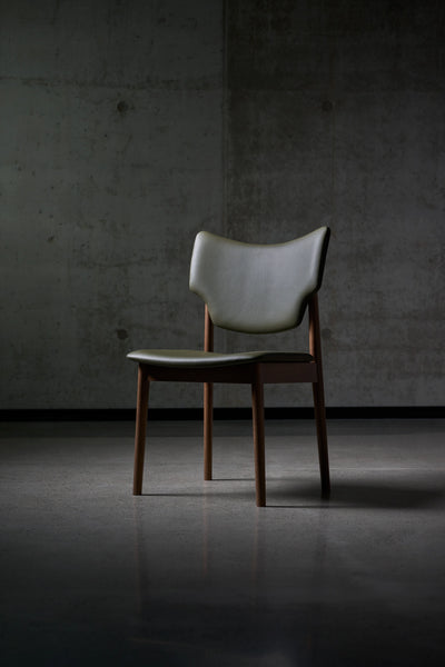 Angled front view of the gray-green Eikund Veng Dining Chair against a concrete wall, showing its sculpted backrest, separate seat, and tapered brown wood legs.