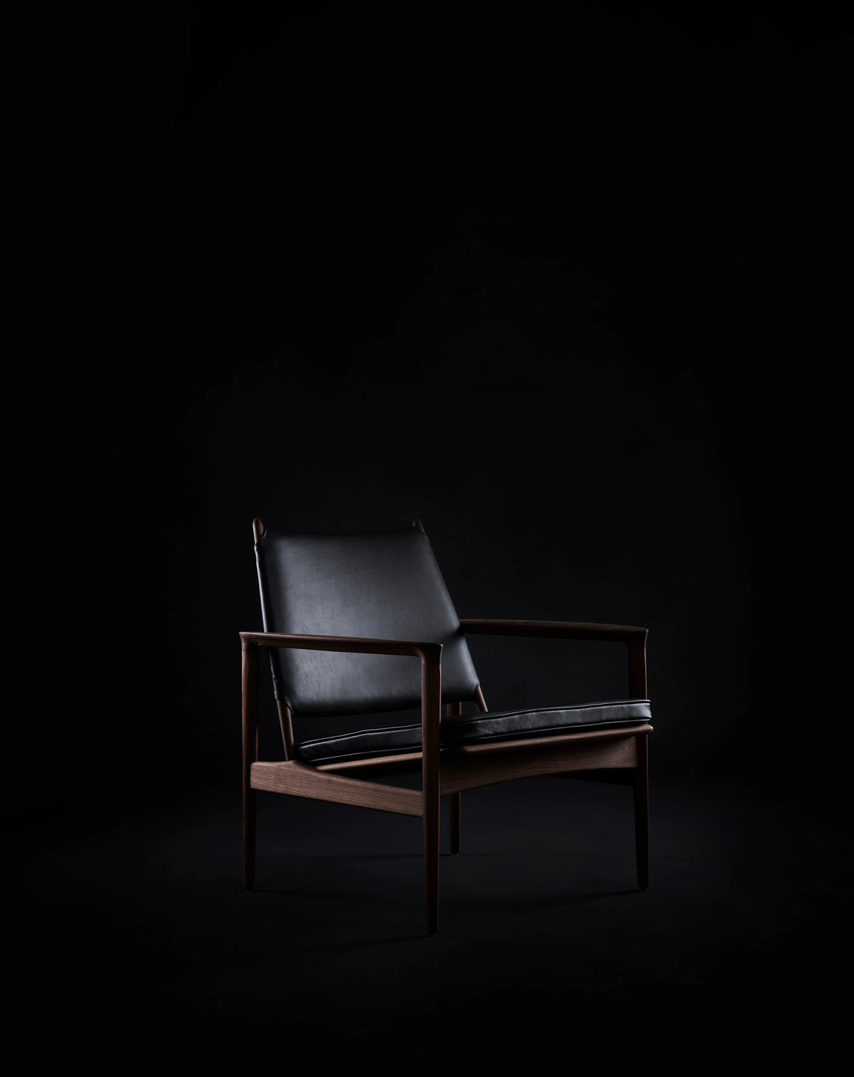 Black leather Eikund Broadway Lounge Chair with a dark wood frame in a dim studio, shown at a three-quarter angle with light highlighting its backrest, armrests, and seat.