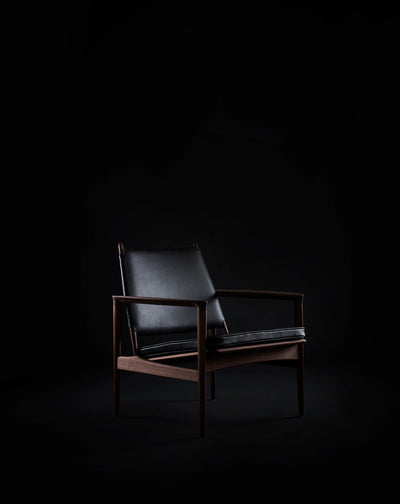 Black leather Eikund Broadway Lounge Chair with a dark wood frame in a dim studio, shown at a three-quarter angle with light highlighting its backrest, armrests, and seat.